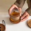 Hands holding a glass of amber liquid above a wooden coaster and iron rack.