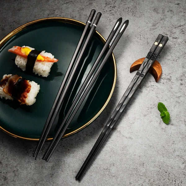 Ein Set schwarzer Edelstahl-Stäbchen liegt auf einem dunkelgrünen Teller mit Sushi und einem Holzstück.