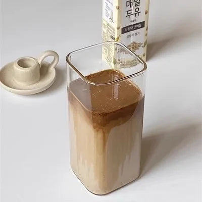 A square heat resistant glass drink cup filled with a light brown beverage, next to a small ceramic saucer and a box.