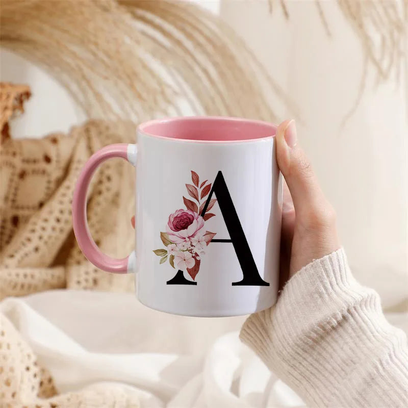 A white coffee mug with a pink handle featuring a large black letter A and floral accents, held by a person.