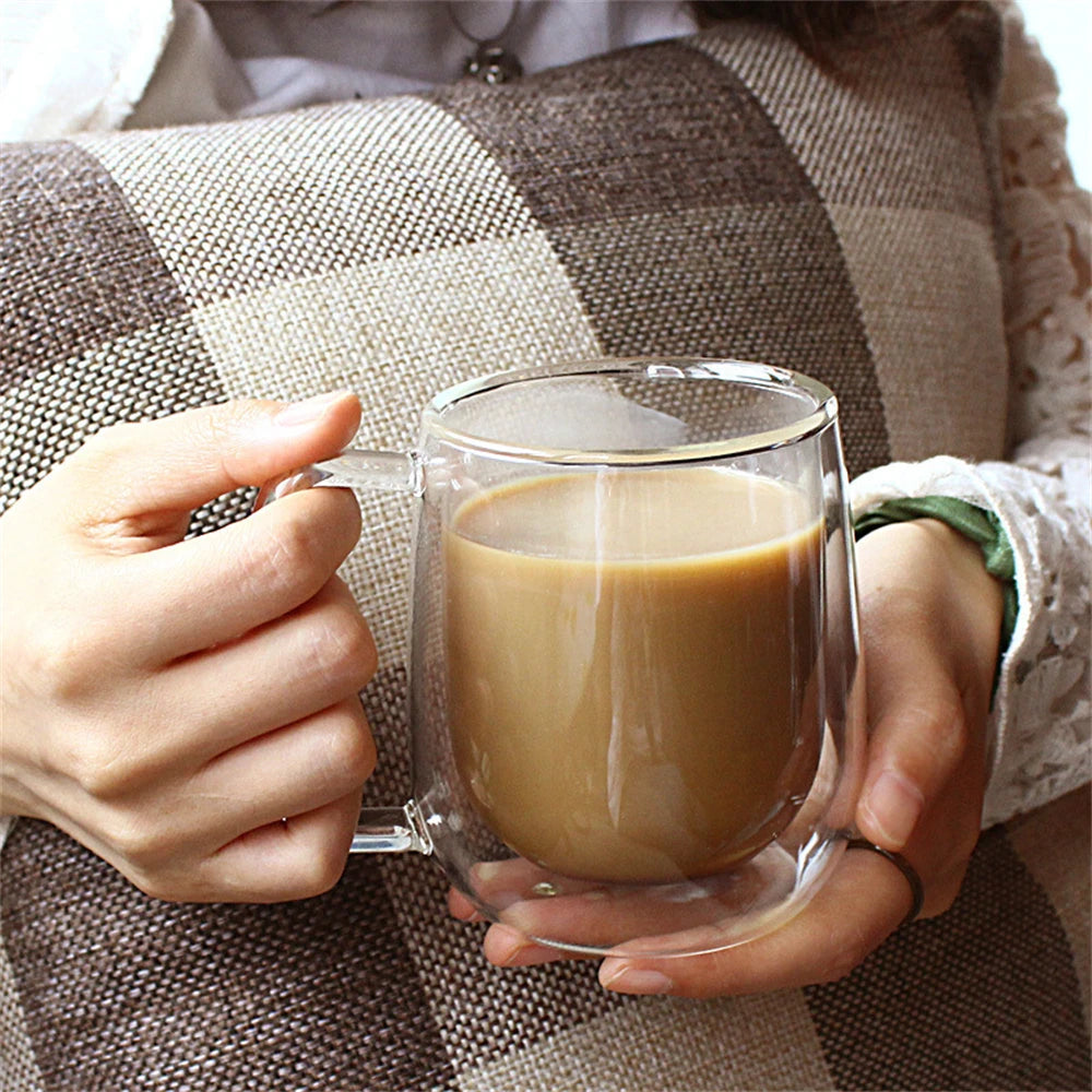 A person holds a double wall insulated glass coffee cup with a handle against a plaid fabric background.