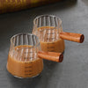 Una taza medidora de espresso de vidrio transparente con mango de madera muestra varias medidas de líquido.