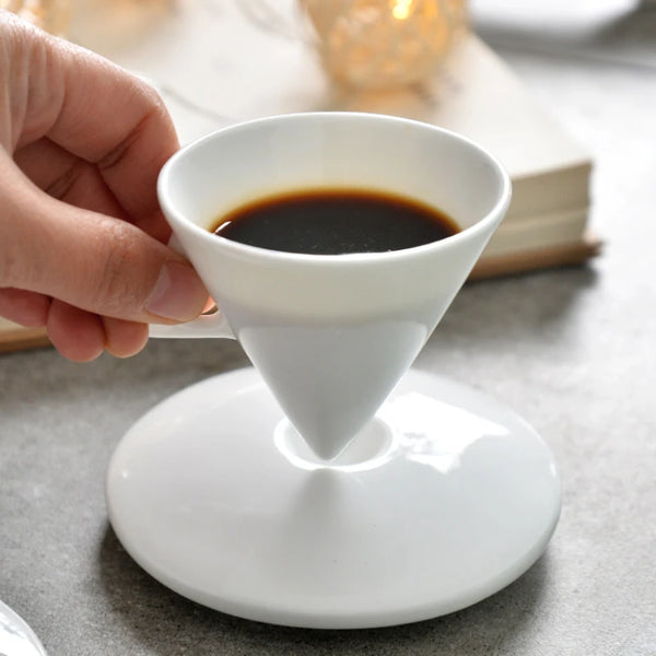 Eine Hand hält einen weißen Keramik-Kaffeefilter mit gebrühtem Kaffee auf einer passenden Untertasse.