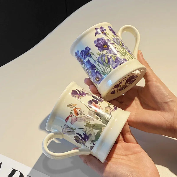 A hand holds a ceramic coffee mug featuring purple and blue floral designs.