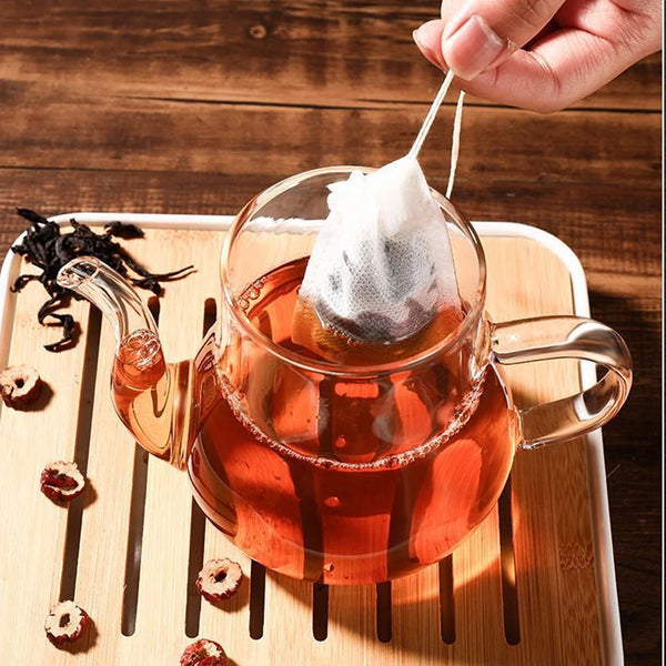A glass teapot filled with amber tea is resting on a wooden tray with tea infuser bags.