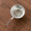 A silver stainless steel tea strainer filter with a wooden handle resting on a brown wooden surface.