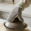 A hand wearing striped baking gloves rests on a ceramic pot or dish.