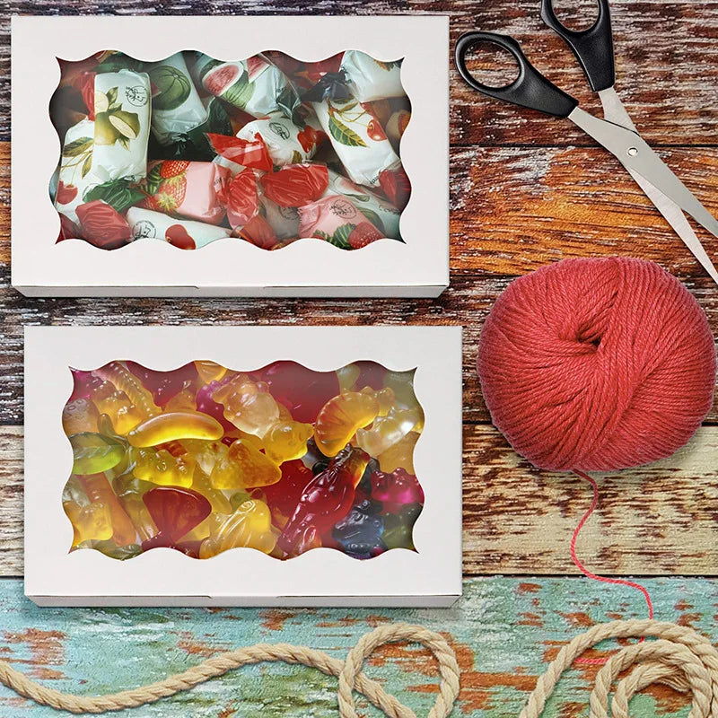 Two decorative kraft paper candy boxes are shown next to a ball of red yarn and scissors on a wooden surface.