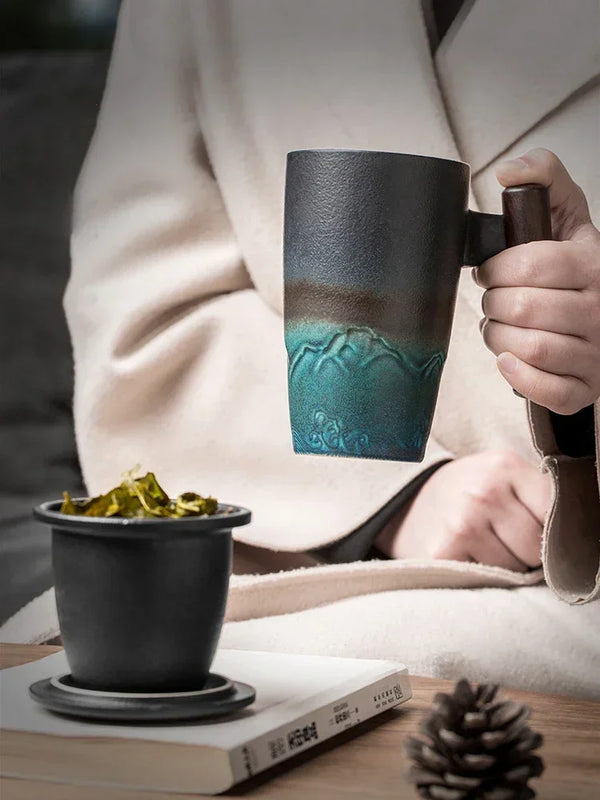 A hand holds a dark ceramic tea mug set with a lid, resting on a book and next to a pine cone.